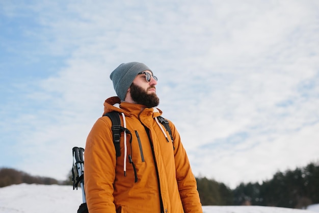 冬の山と森を背景にサングラスと暖かい服を着たバックパックを持つひげを生やした男 ハイキングとアクティブなライフスタイルのコンセプト