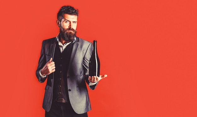 Bearded man with a bottle champagne of and glass. the person\
holds a red wine bottle in a hand. stylish male in tuxedo, suit,\
jacket.