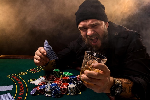 Foto uomo barbuto con sigaro e bicchiere seduto al tavolo da poker in un casinò. gioco d'azzardo, carte da gioco e roulette.