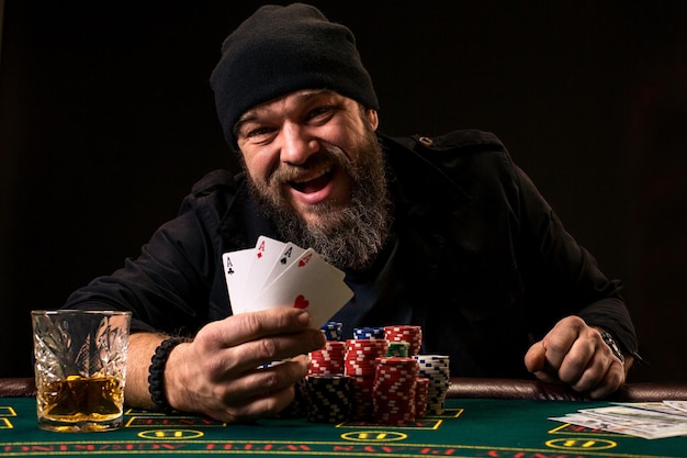 Bearded man with cigar and glass sitting at poker table and screaming isolated on black