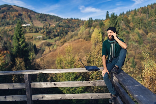 Bearded man with laptop sits on railing and talks on phone Freelancer hipster on vacation in mountains