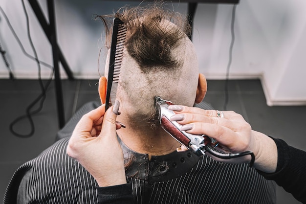 Парикмахер стрижет бородатого мужчину с ирокезом, сидя в кресле в парикмахерской. Девушка-парикмахер делает ирокез парню