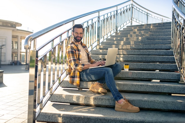 Бородатый мужчина работает на ноутбуке на лестнице