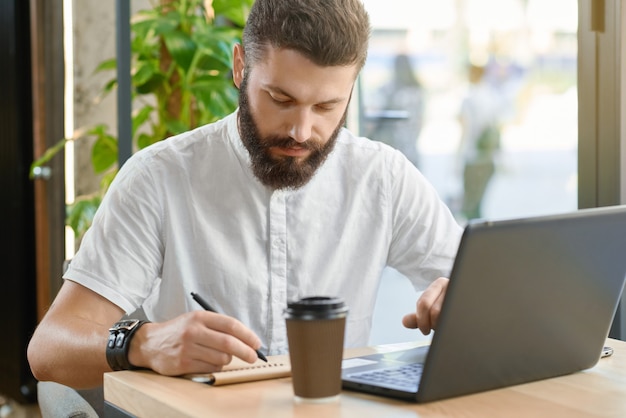 수염 난된 남자 메모 작성, 창 근처에 앉아 노트북으로 작업.