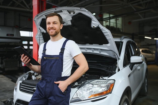 Bearded mechanic in overalls standing in garage of a car salon and holding tablet He is about to diagnostic breakdown