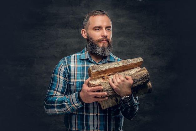 A bearded middle age male dressed in plaid flannel shirt holds firewoods over a dark background.