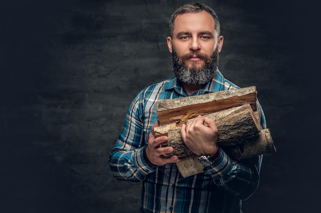 A bearded middle age male dressed in plaid flannel shirt holds firewoods over a dark background.