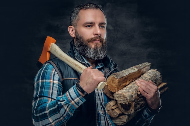 Bearded middle age male holds an axe and firewoods over dark grey background.