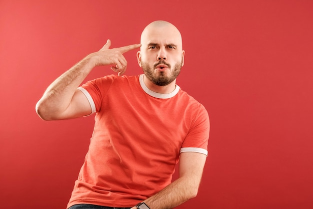 A bearded middle-aged man in a red T-shirt on a red background shows his hand a gun to his temple. Isolated.