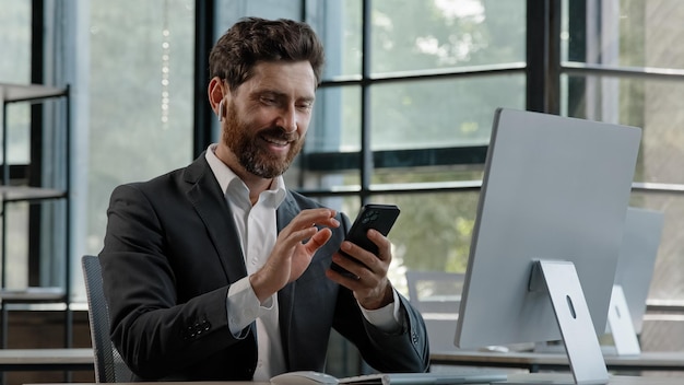 Bearded middleaged business man in office sitting table using phone mobile audio app service to