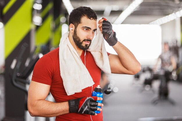 ひげを生やした筋肉の男は、ジムの勤勉な休息でタオルとボトルと赤いTシャツを着ています