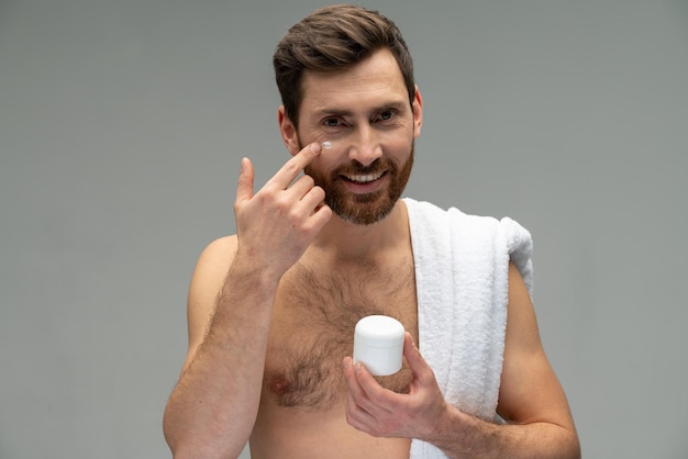 Bearded naked young man with perfect skin applying moisturiser\
facial cream isolated on grey background studio portrait skin care\
healthcare cosmetic procedures concept