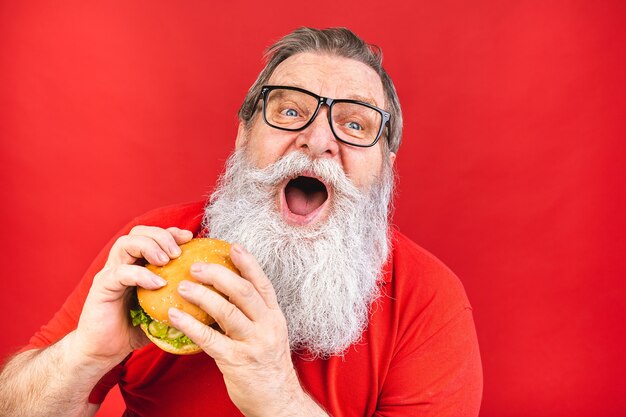 bearded old man with glasses in red tshirt eating a hamburger isolated on red background