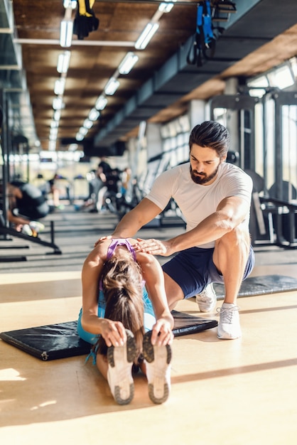 Barbuto personal trainer aiutando la donna ad allungare. donna seduta sulla stuoia.