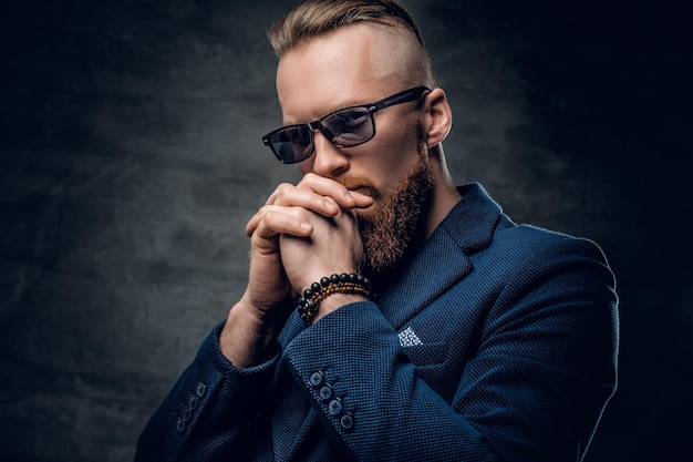 Bearded redhead male dressed in a blue suit and sunglasses over dark grey background.