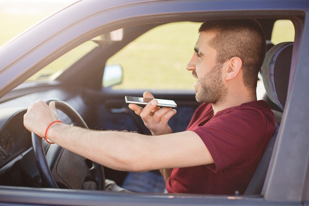 Bearded satisfied male driver drives car, speaks via smart phone, dressed casually, happy to hear friend, focused on road