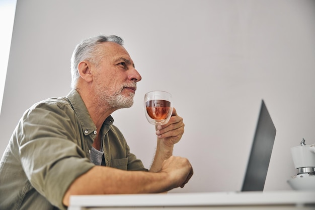 Signore anziano barbuto che beve tè mentre lavora al computer portatile