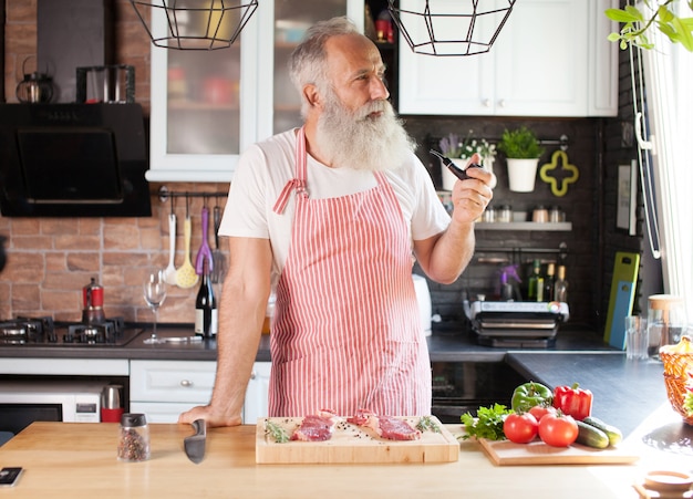 ひげを生やした年配の男性が彼のキッチンでステーキを調理します。