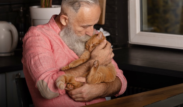 家でかわいい猫を保持しているひげを生やした年配の男性。