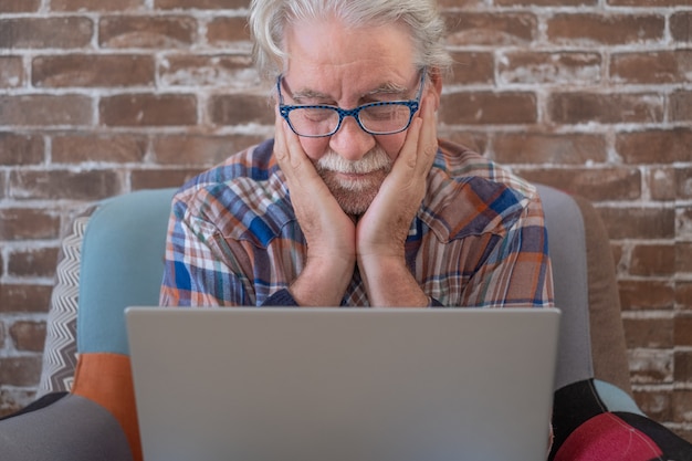Bearded senior man sitting at home on armchair using laptop computer. Brick wall in background