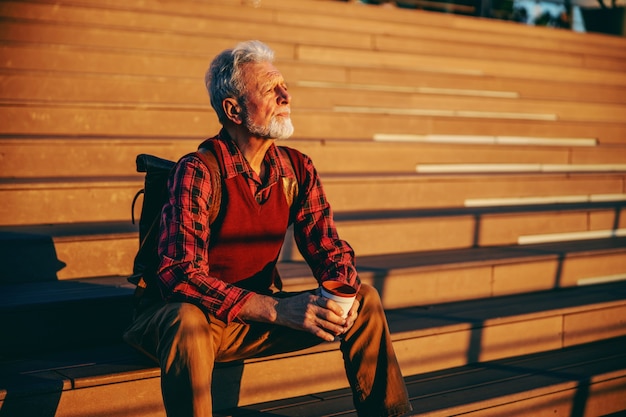 Bearded senior man sitting on the stairs outdoors, looking at something and drinking coffee to go