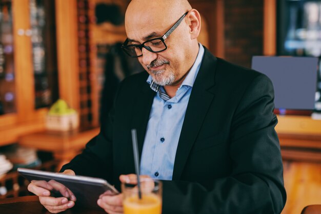 Bearded senior in suit using tablet while sitting in cafeteria.