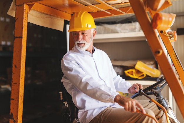 Bearded senior worker in uniform and with protective helmet on head driving forklift in storage.