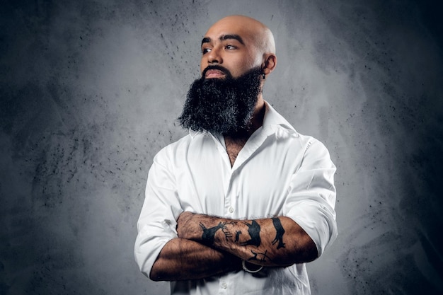 Bearded shaved head Arabic male with tattoos on his crossed arms, dressed in a white shirt posing over grey background.