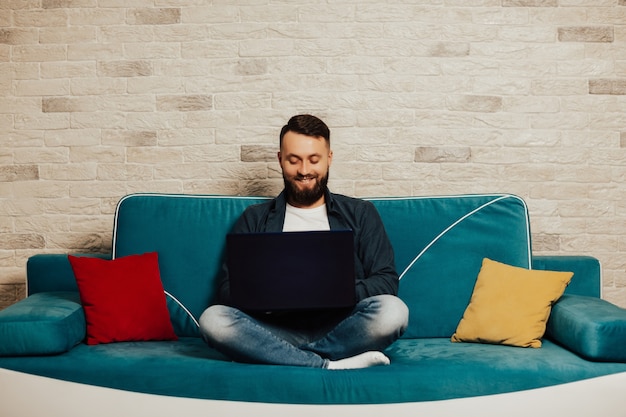 Bearded smiling man looking at laptop screen, while sitting on turquoise sofa at home.