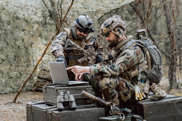 Bearded soldiers in uniform sit on military transport crates analyze data on a laptop and work out