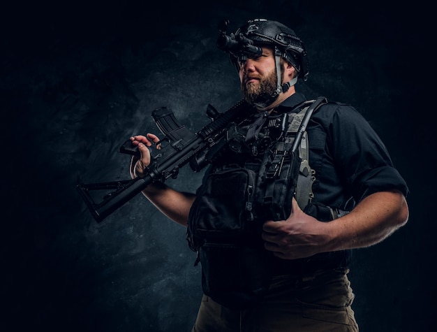 Bearded special forces soldier or private military contractor holding an assault rifle and observes the surroundings in night vision goggles. Studio photo against a dark textured wall