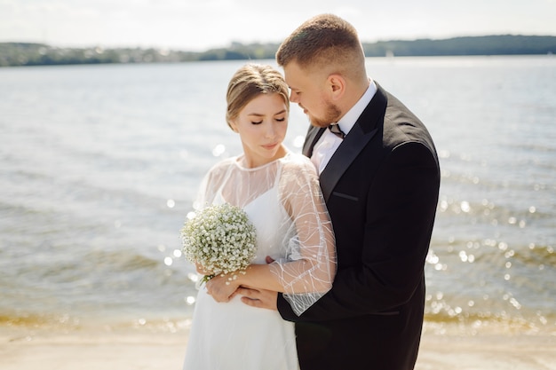 Uno sposo barbuto ed elegante in abito e una bella sposa bionda in abito bianco con un bouquet in mano sono in piedi e si abbracciano nella natura nella foresta di pini.