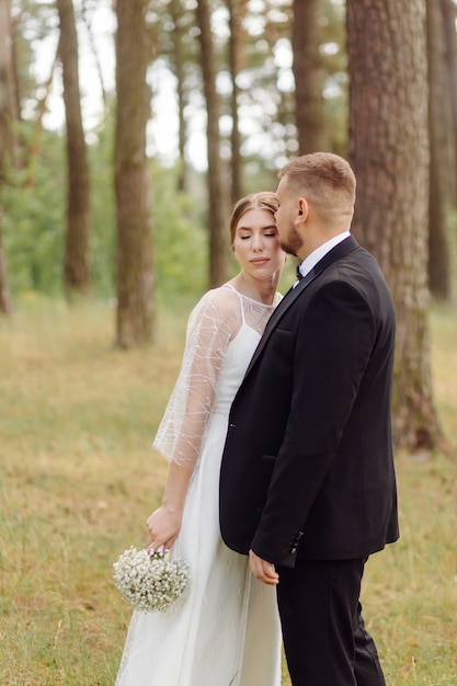 A bearded, stylish groom in a suit and a beautiful blonde bride in a white dress with a bouquet in her hands are standing and hugging in nature in the pine forest.