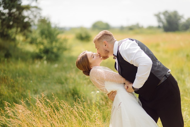 A bearded, stylish groom in a suit and a beautiful blonde bride in a white dress with a bouquet in her hands are standing and hugging in nature in the pine forest.