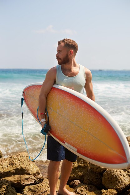 Bearded surfer with a surfboard is walking on the beach and watching for the waves on the spot