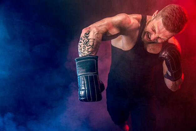Photo bearded tattooed sportsman muay thai boxer in black undershirt and boxing gloves fighting on dark background with smoke. sport concept.