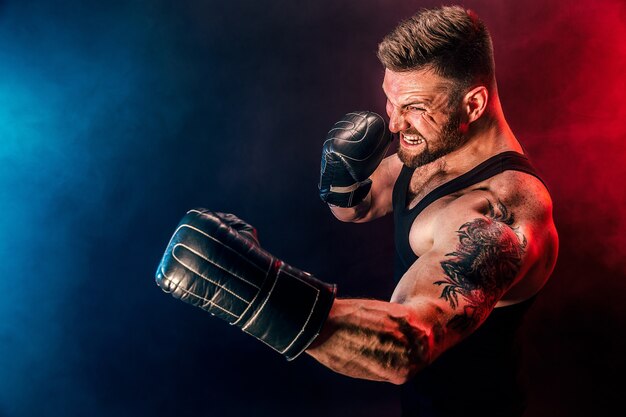 Bearded tattooed sportsman muay thai boxer in black undershirt and boxing gloves fighting on dark background with smoke. Sport concept.