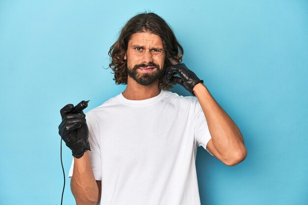 Photo bearded tattooist with tattoo machine at work covering ears with hands