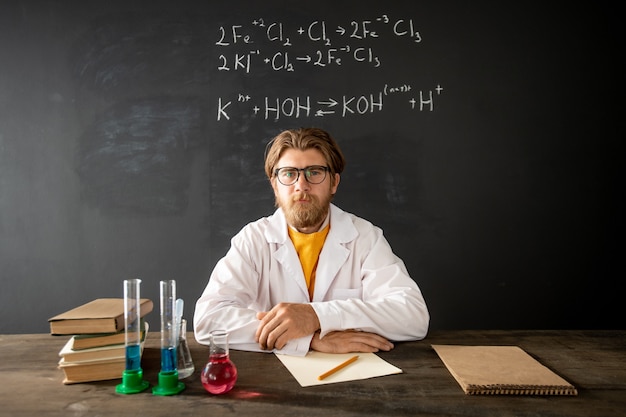 Bearded teacher of chemistry looking at tube with pink liquid substance while sitting by table in front of smartphone camera at lesson