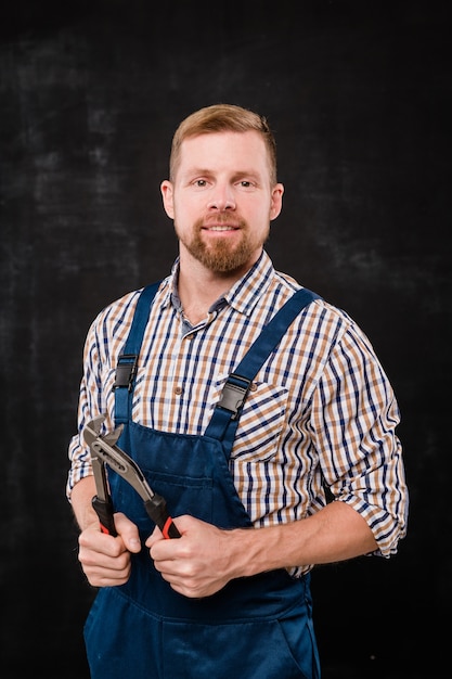 Bearded technician or repairman in shirt and overalls using handtool in isolation over black background