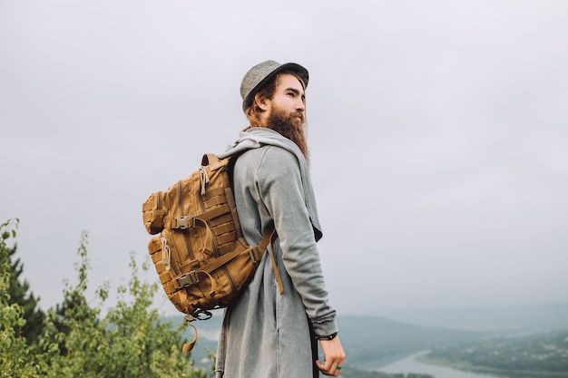 Bearded tourist man in a grey hat with a backpack is standing and watching the landscape