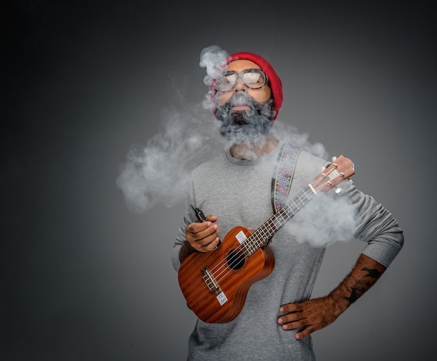 Bearded unformal male holding acoustic small guitar and smoking a cigarette.