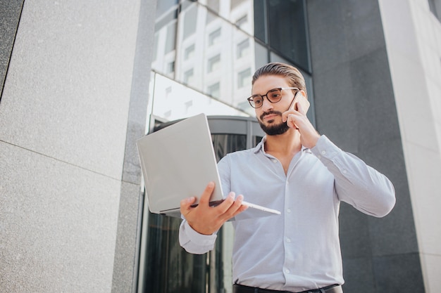 L'uomo d'affari barbuto e giovane sta alla costruzione e parla sul telefono. tiene il portatile con una mano e guarda il suo schermo. guy è concentrato.