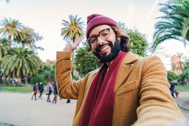 Bearded young caucasian male taking a selfie portrait with a smartphone in a park outdoors wearing a beanie hat glasses and scarf in autumn season Close up photo of a happy man in a tourism trip