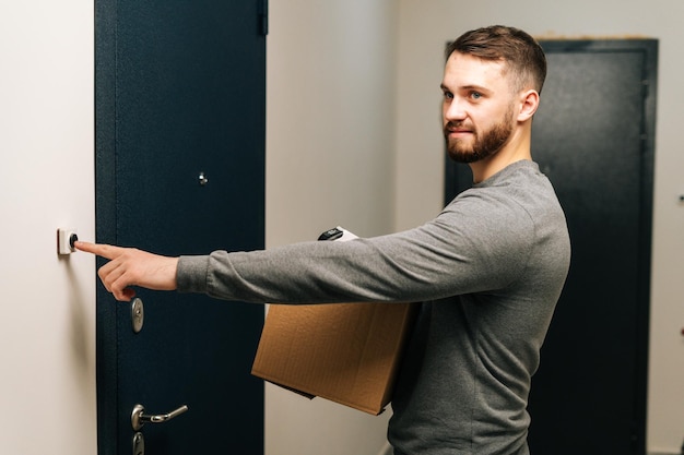 Bearded young delivery man holding in hand cardboard box with parcel and ringing doorbell of customer apartment