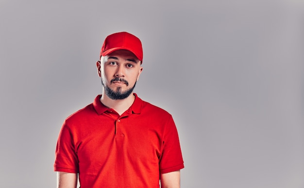 Bearded young delivery man in red t-shirt and cap isolated on gray background. Home delivery service.