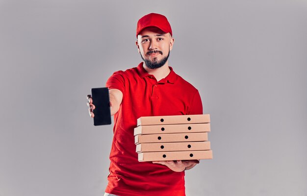 Il giovane fattorino barbuto in maglietta rossa e berretto con scatole per pizza mostra lo schermo vuoto dello smartphone isolato su sfondo grigio. consegna a domicilio veloce.