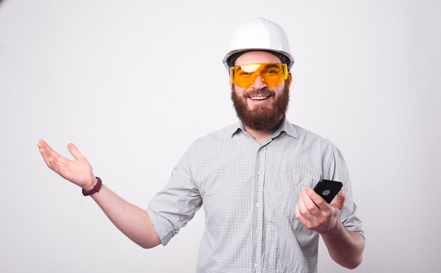 A bearded young engineer is holding a smartphone and smiling at the camera is wearing a helmet and protective glasses near a white wall