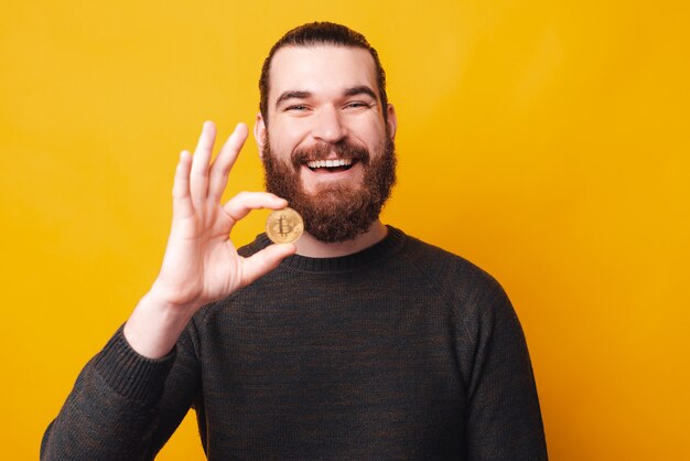 ひげを生やした若い幸せな男は笑顔でビットコインを保持しています