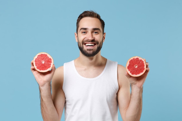 Бородатый молодой человек 20 лет в белой рубашке держит в руке половину грейпфрута, изолированного на синем пастельном фоне, студийный портрет. Концепция косметических процедур по уходу за кожей. Макет копировального пространства